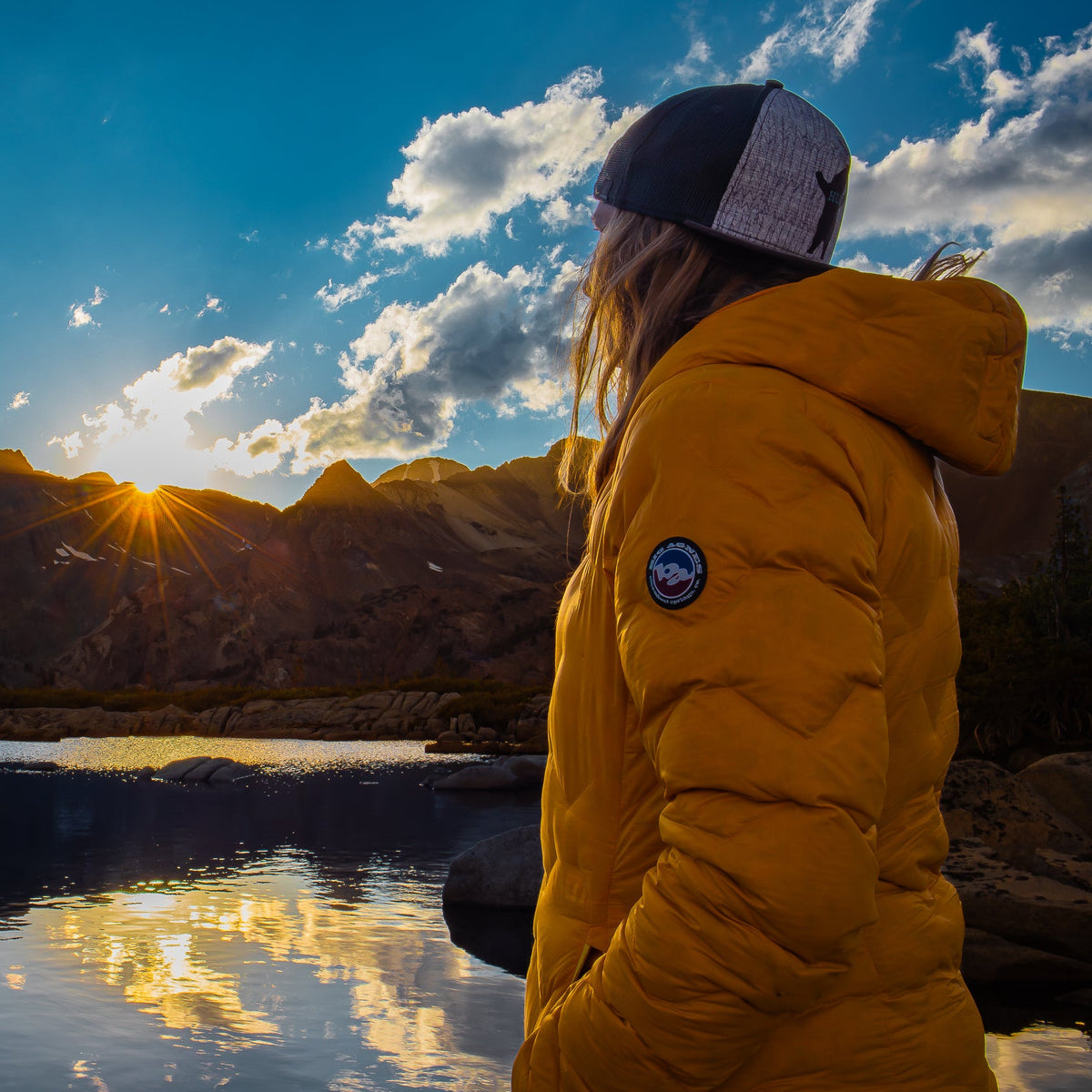 Person in a yellow jacket and cap standing by a body of water with mountains in the background