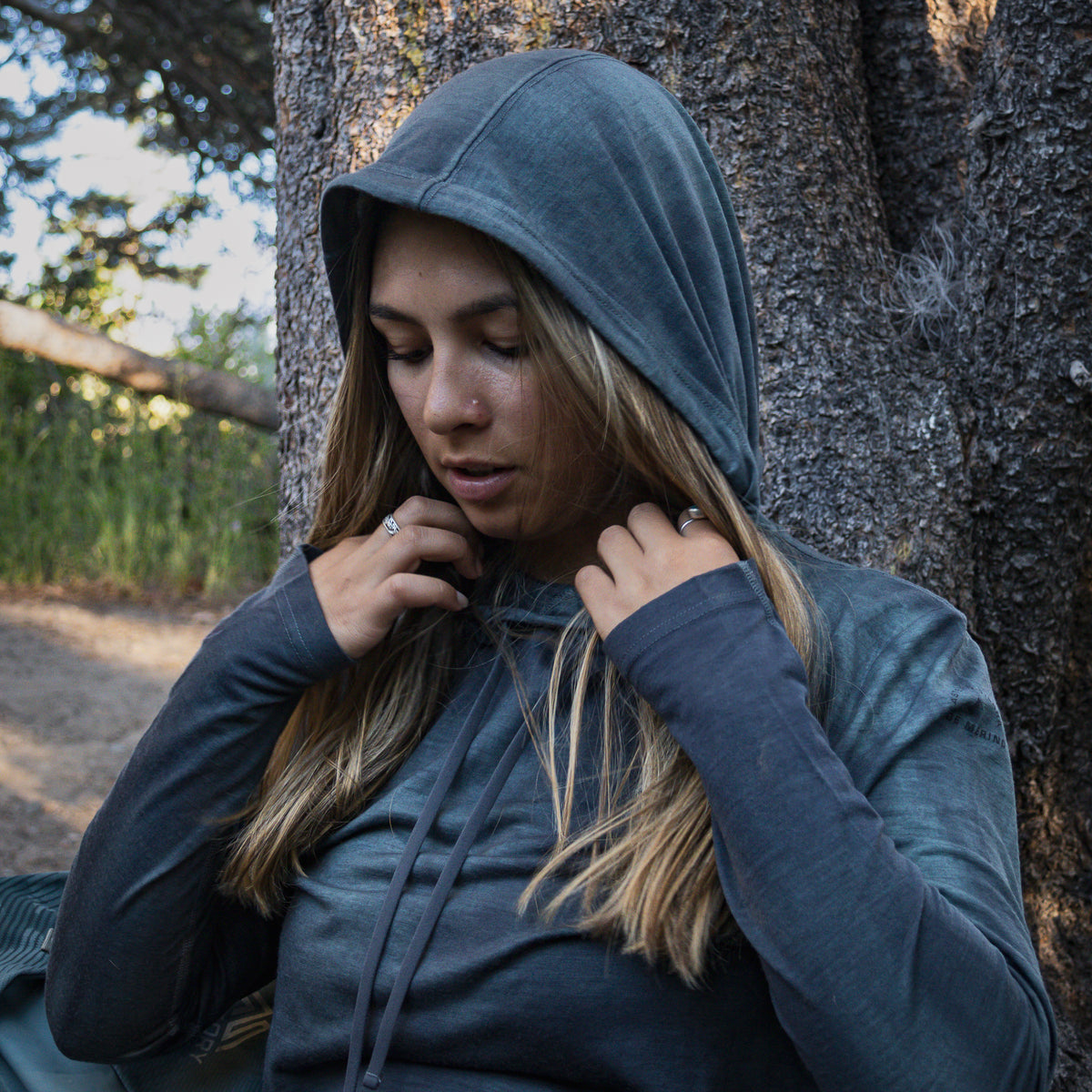 Woman in a blue hoodie sitting against a tree in a natural setting