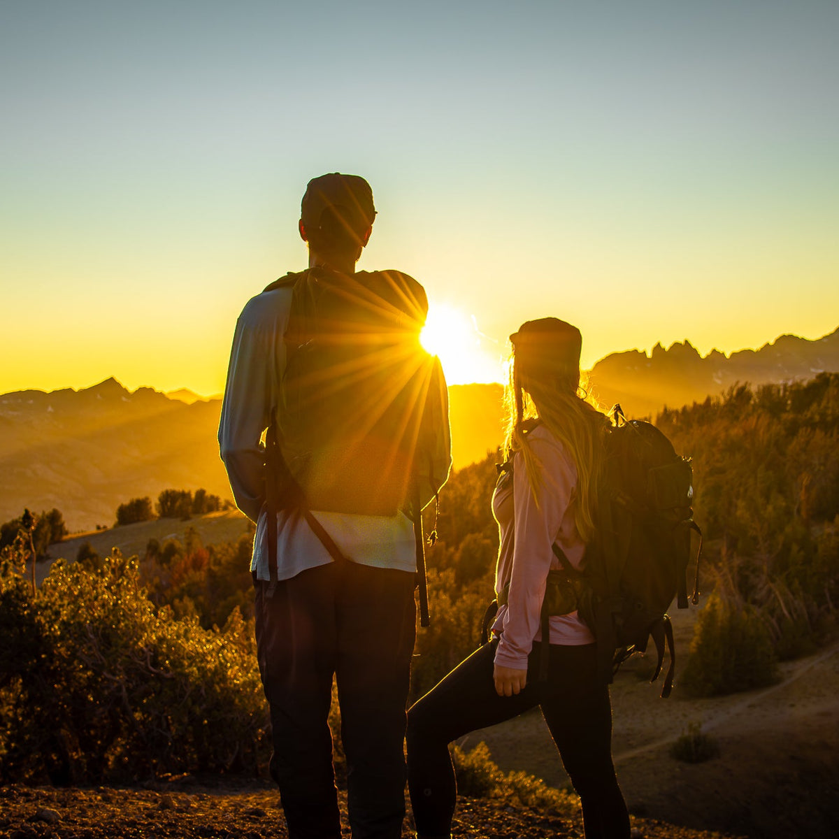 Two people with backpacks standing on a trail at sunset with mountains in the background