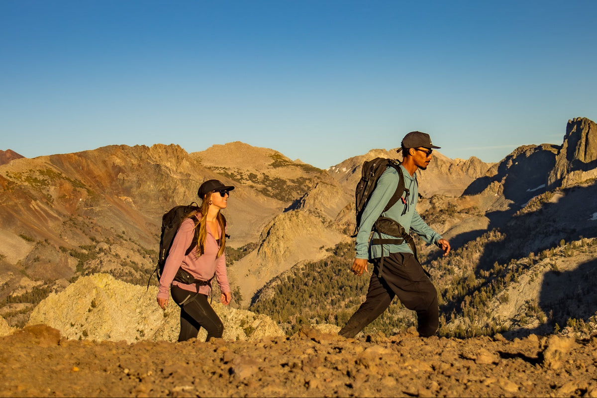 Two hikers with backpacks walking on a mountain trail under a clear blue sky.
